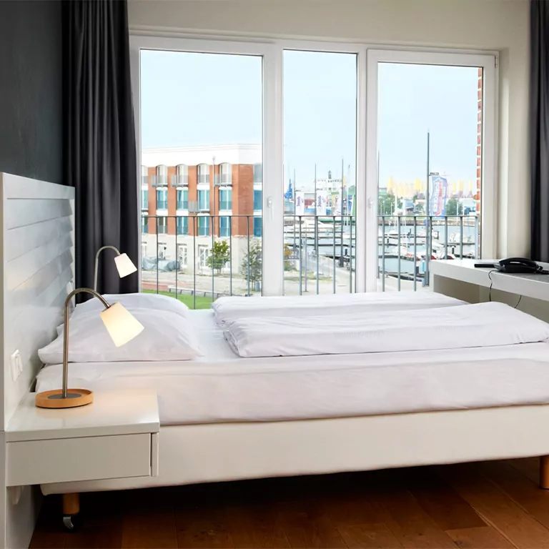 Modernes Hotelzimmer mit Doppelbett, Blick auf den Hafen durch große Fenster, stilvolle Nachttischlampen im ATLANTIC Hotel Sail City.