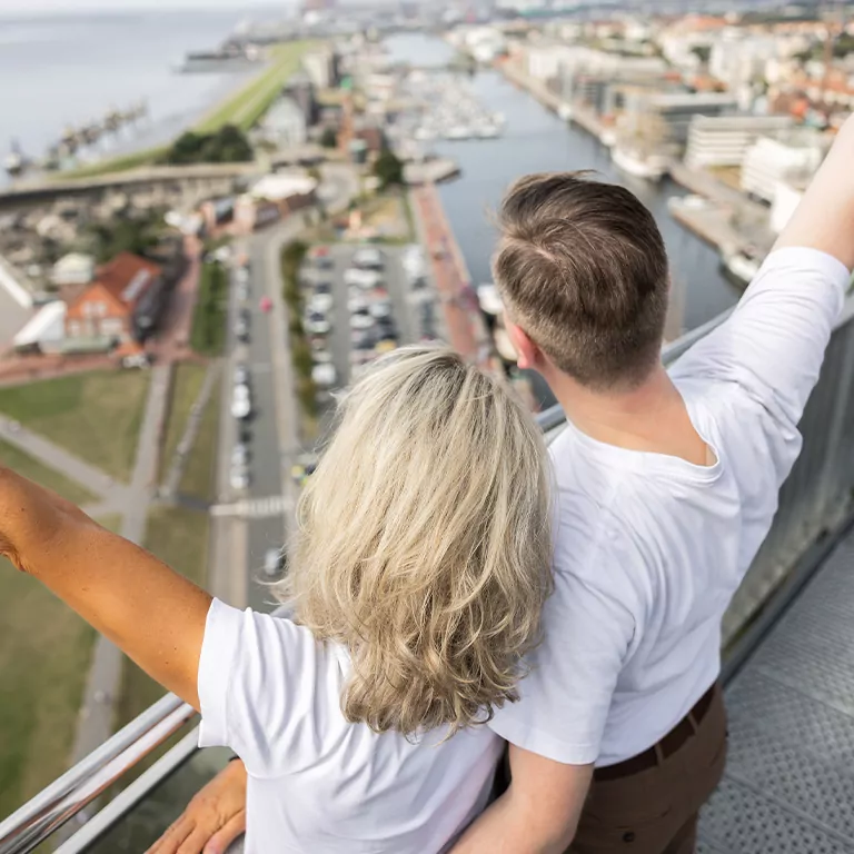 Paar genießt den Ausblick von einer Aussichtsplattform mit Blick auf eine Küstenstadt und den Hafen.