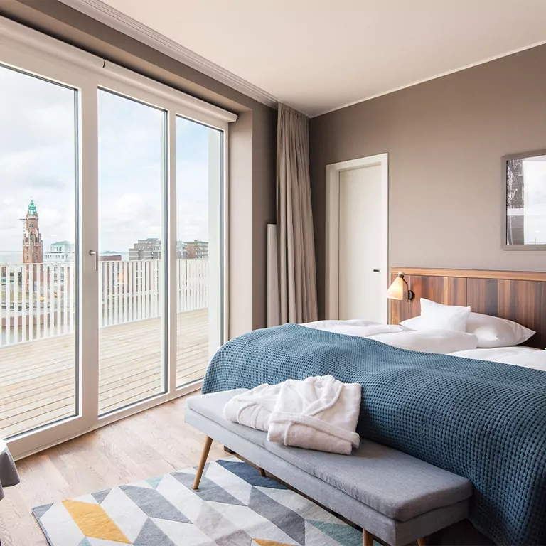 Helles Hotelzimmer mit Doppelbett, großer Fensterfront und Balkon mit Stadtblick.