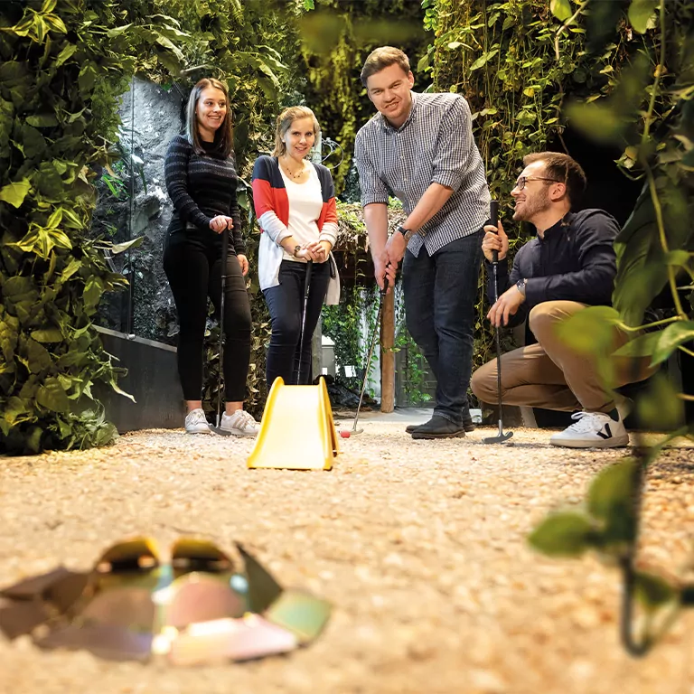 Vier Personen spielen Minigolf in einem üppig begrünten, indoor Minigolf-Parcours.