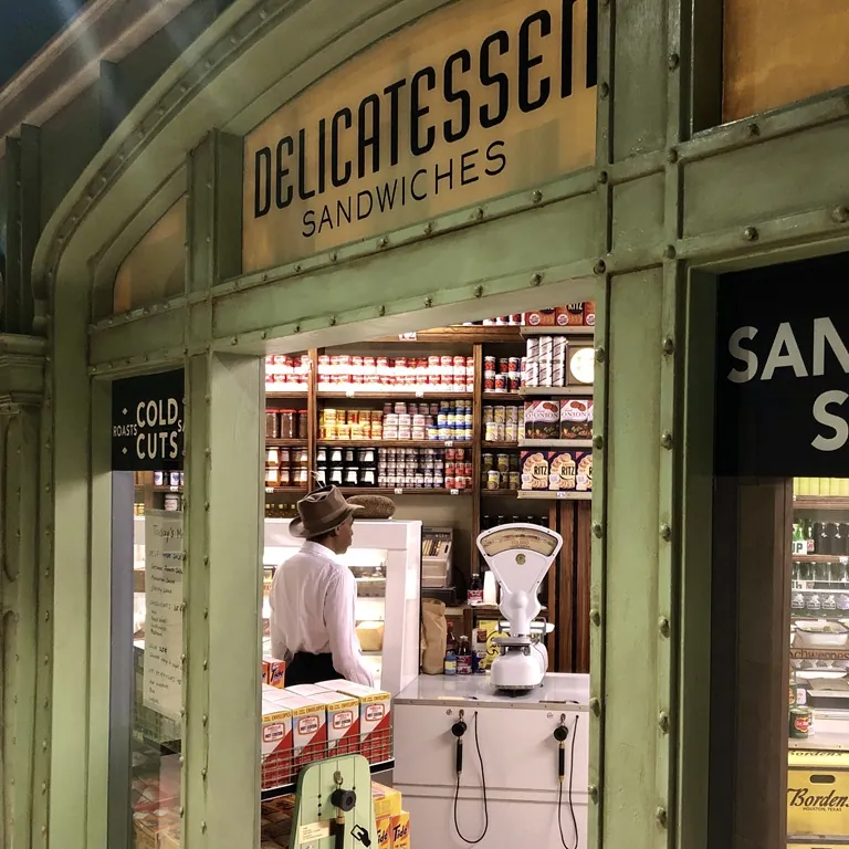 Deutsches Auswandererhaus Austellunsraum - Delikatessengeschäft mit Sandwiches, Regale voller Produkte, ein Verkäufer steht hinter der Theke, klassische Waage im Vordergrund.