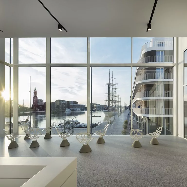 Moderner Raum im deutschen Auswandererhaus mit großen Fenstern, Blick auf Hafen mit Segelschiff und Gebäude, stilvolle Stühle im Vordergrund.
